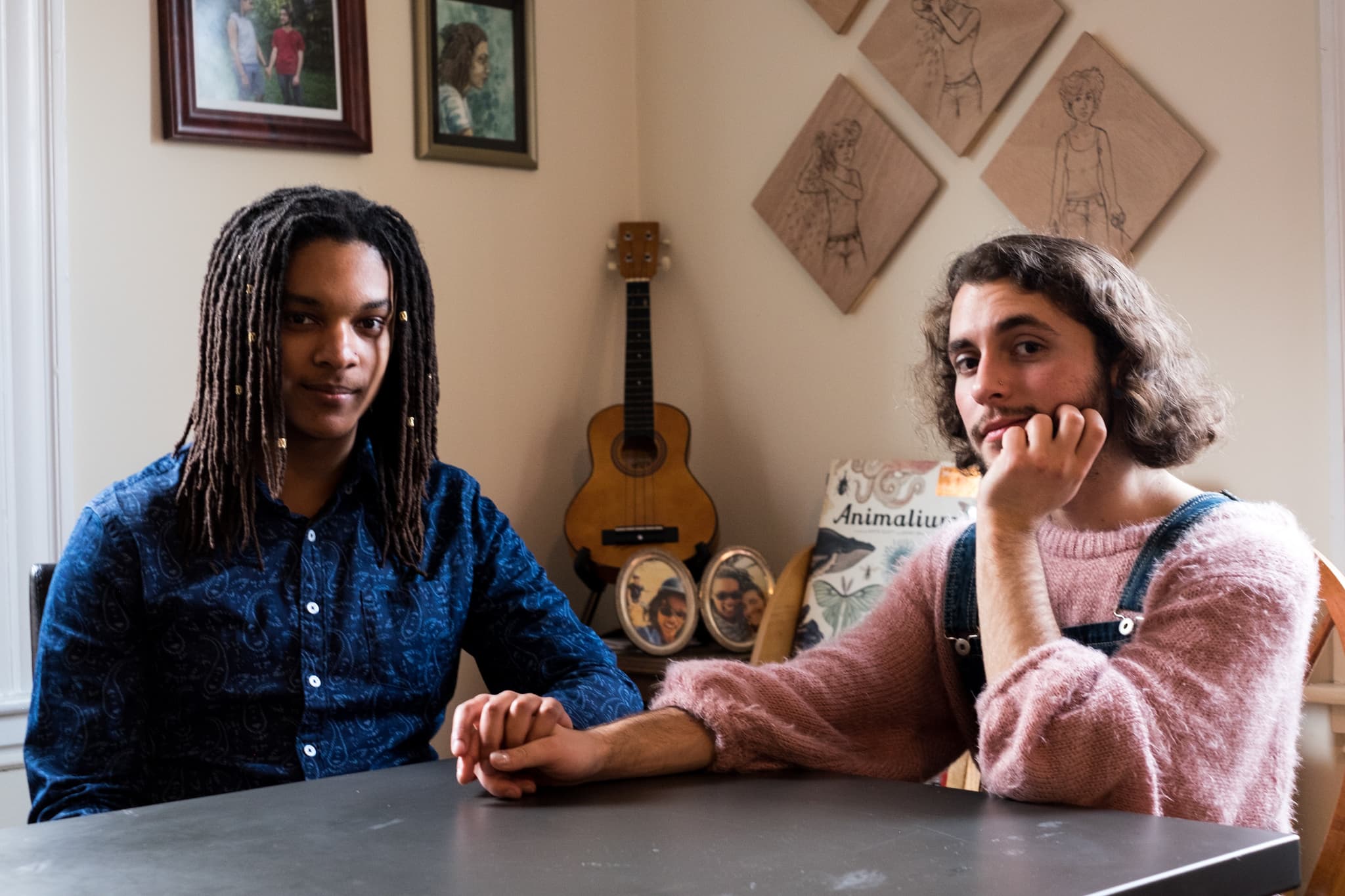 Two figures seated at a table hold hands, a guitar appearing by the wall behind them.